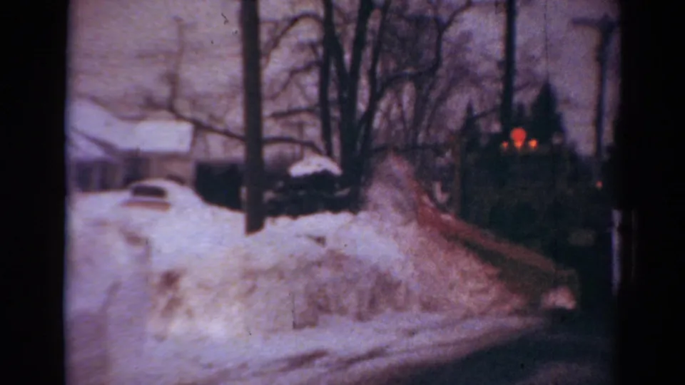 1954 Toledo frame with a snowplow truck moving along a residential road lined by snowbanks, trees, and houses.