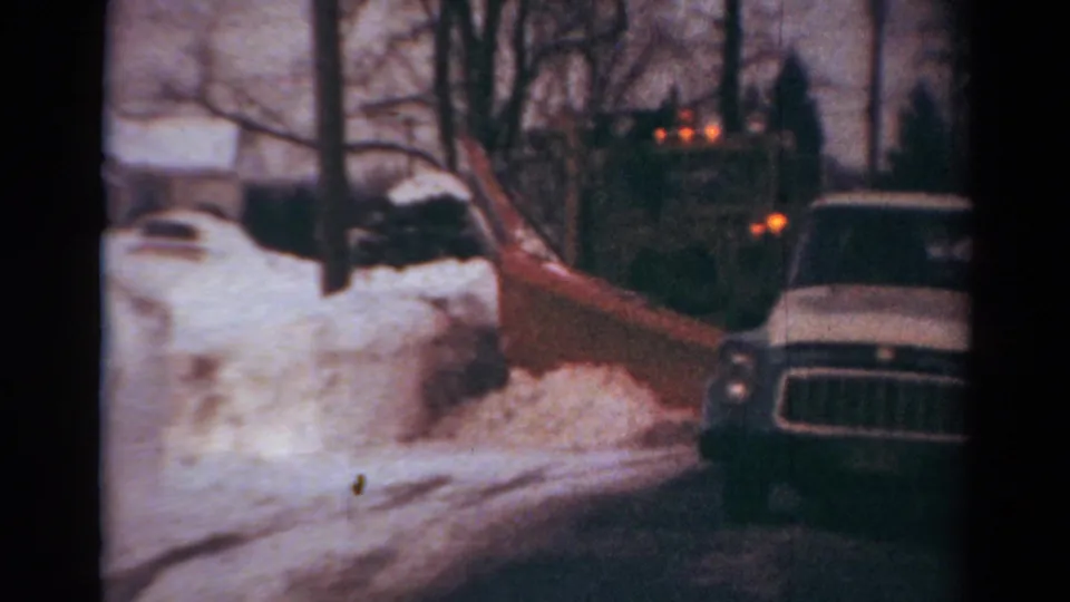1954 Toledo home-movie frame showing the snowplow close to the camera, with a parked vehicle and deep snowbanks nearby.