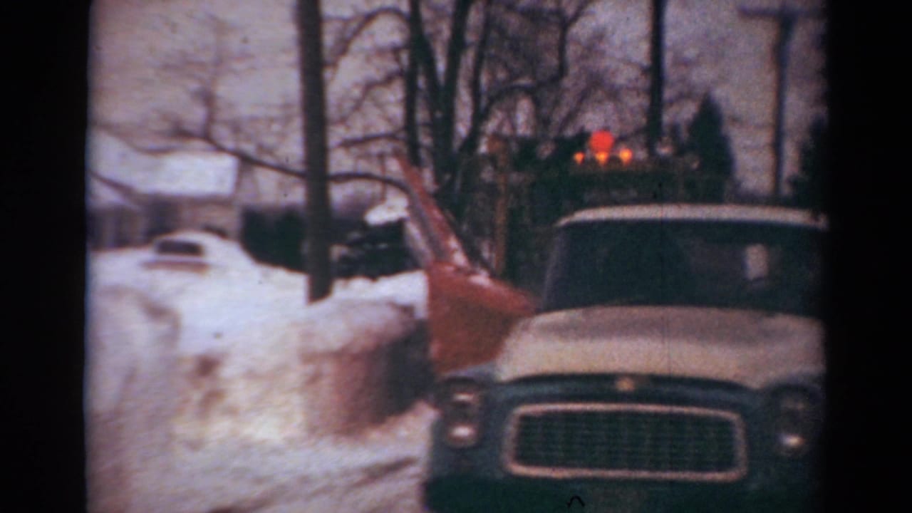 Snowplow truck faces the camera on a snowy residential street with a white car at left.