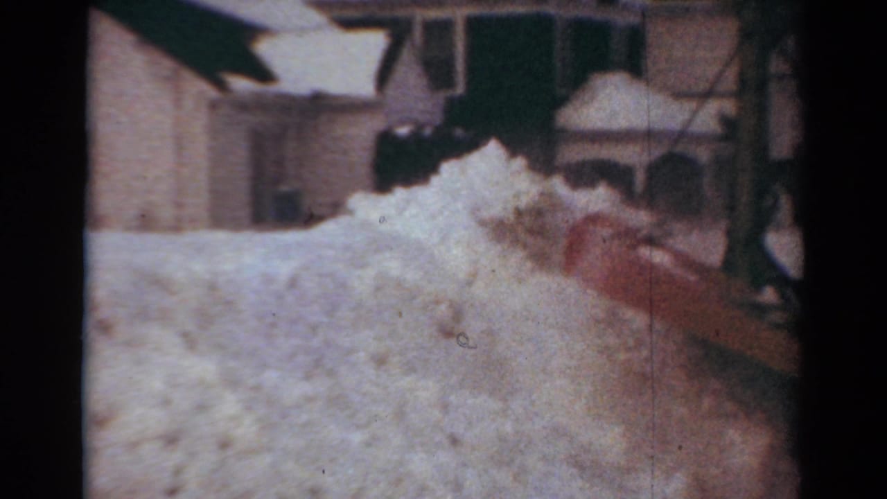 Red plow blade pushes a high mound of snow near houses.