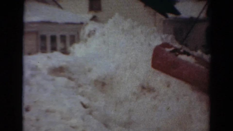 The plow blade pushes a spray of snow across the frame, with residential windows visible in the background.