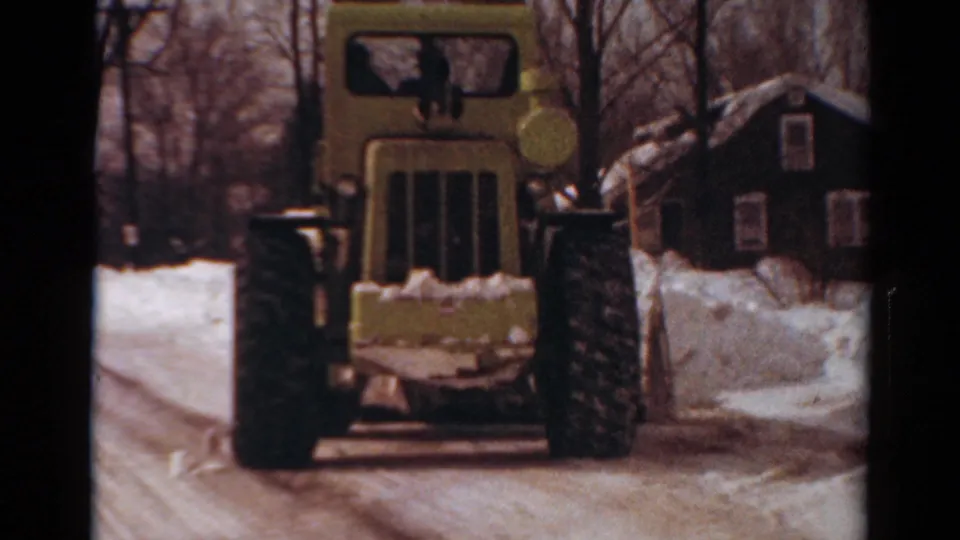 1954 Toledo home-movie frame showing a yellow wheeled bulldozer or loader approaching on a snow-covered residential street.