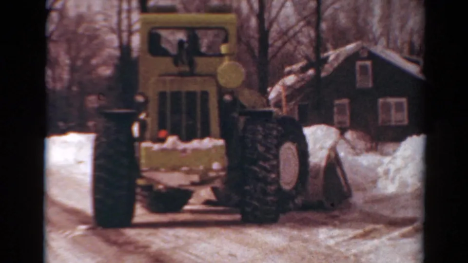 Yellow snow-removal vehicle advances on a snowy Toledo residential road, with a dark house and bare trees in the background.