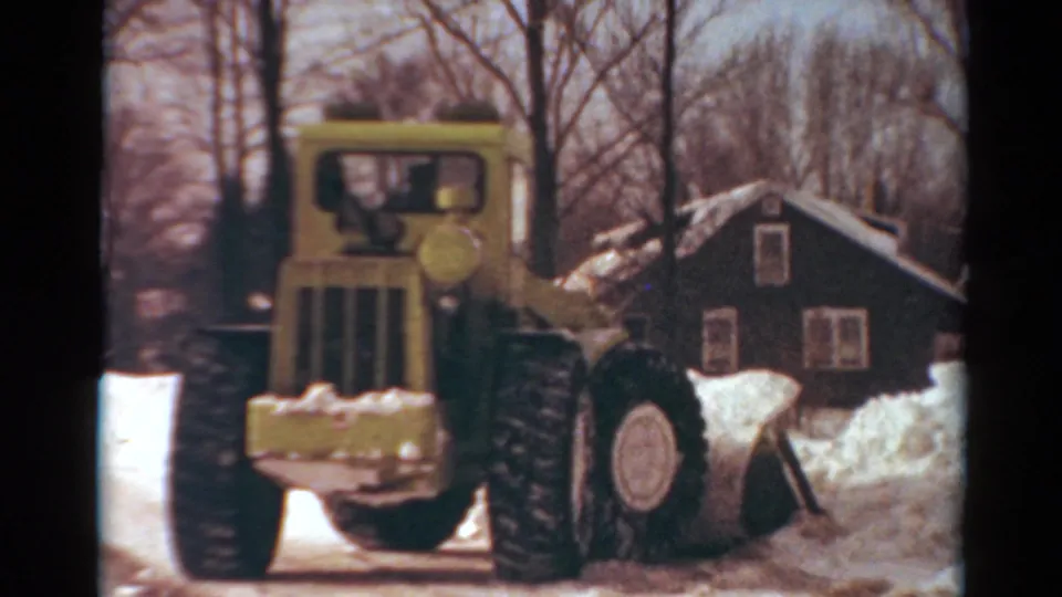 Late frame of the yellow snow-clearing machine on a snowy residential street, with a dark house and winter trees behind it.