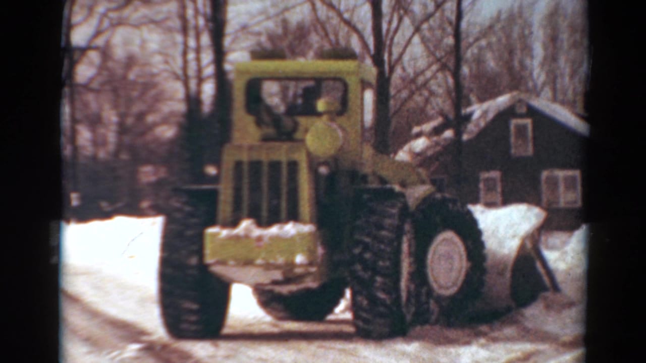 1954 Toledo home-movie frame showing a yellow snow plow approaching through roadside snow near houses and leafless trees.