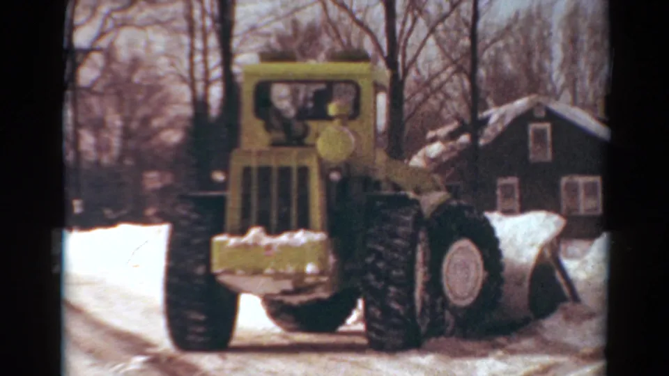 1954 Toledo home-movie frame showing a yellow snow plow approaching through roadside snow near houses and leafless trees.
