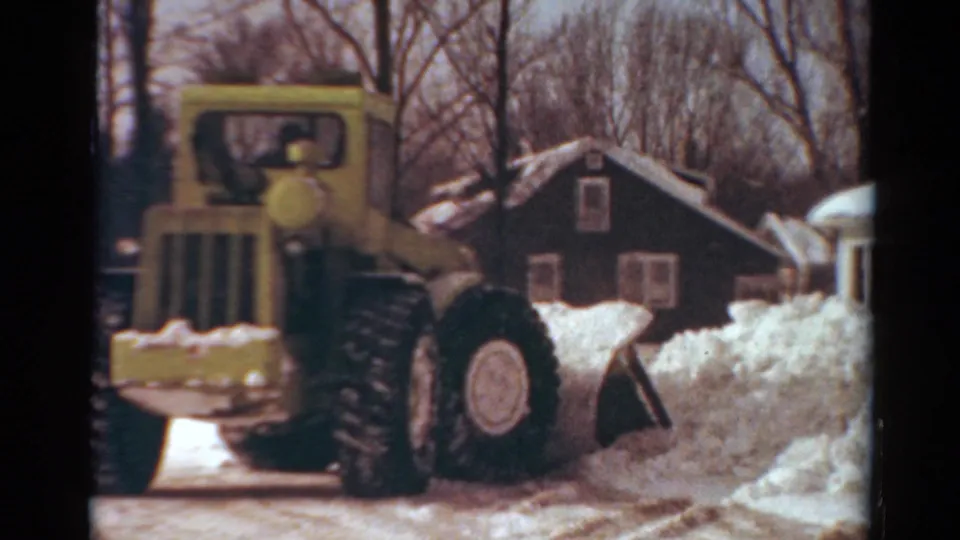 1954 Toledo frame with the yellow plow angled toward piled snow along a residential street beside a dark house.