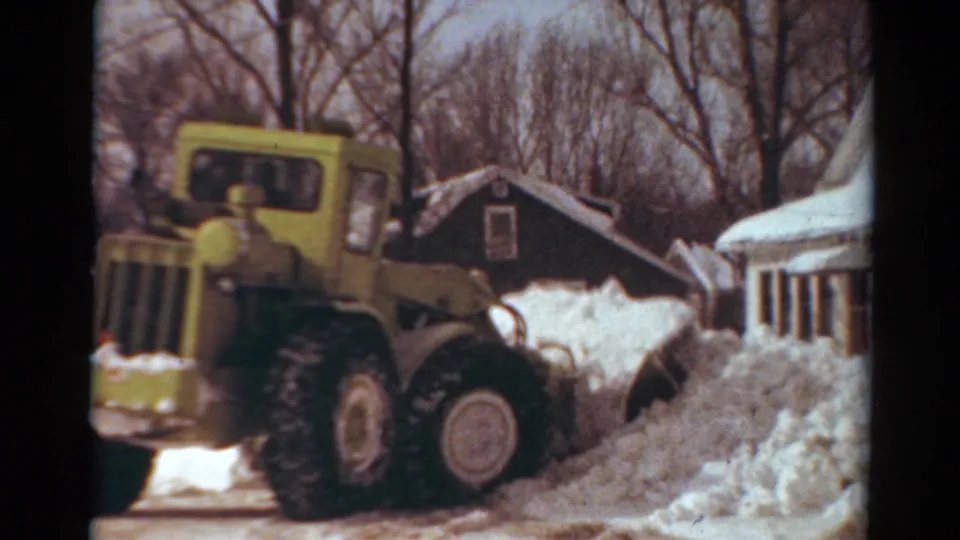 1954 Toledo frame showing the yellow snow plow continuing along the residential street with piled snow beside nearby houses.
