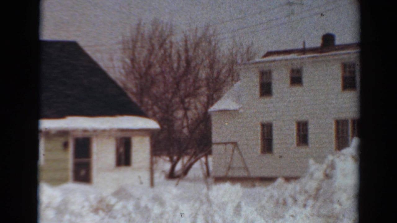 1954 Toledo home-movie frame showing snow piled across a yard beside white residential buildings, bare trees, utility lines, and a swing-set frame.