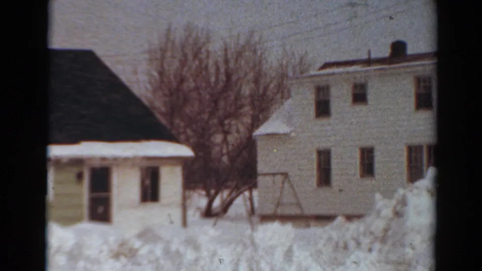 1954 Toledo home-movie frame showing snow piled across a yard beside white residential buildings, bare trees, utility lines, and a swing-set frame.