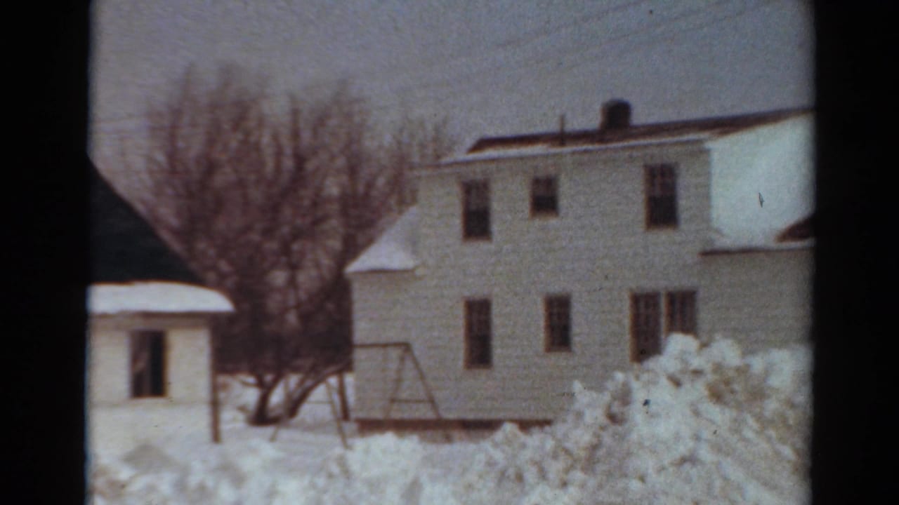 White two-story house and nearby structure behind deep snowbanks in a winter yard.