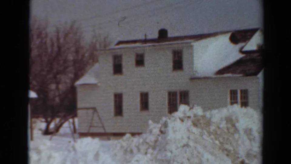 1954 Toledo home-movie frame during the pan, showing the side of a white house, a bare tree, a swing-set frame, and uneven snowbanks across the yard.