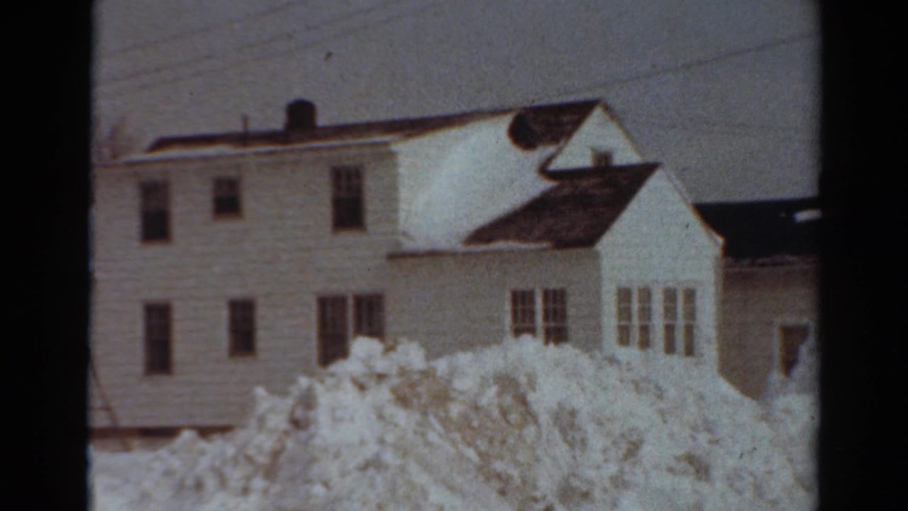 Snow-covered white houses behind high roadside snowbanks under a gray sky.