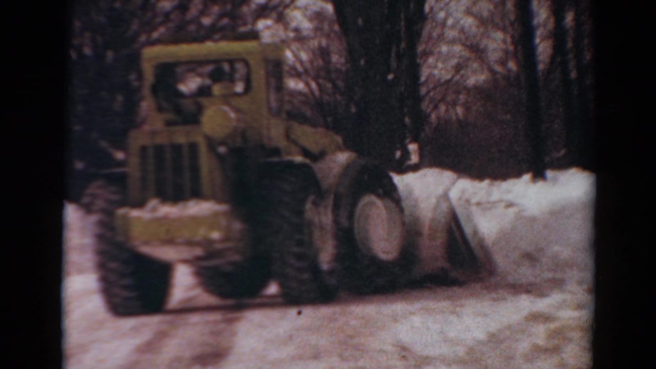 1954 Toledo home-movie frame showing a yellow tractor with front plow moving through deep snow near bare trees.