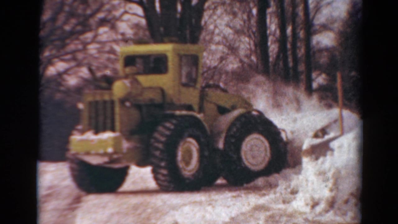 The yellow tractor works beside a tall snowbank in a residential Toledo winter scene, with the driver faintly visible inside the cab.