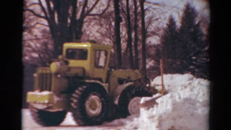 Close home-movie view of the yellow tractor and plow working through snow in the 1954 Toledo residential setting.