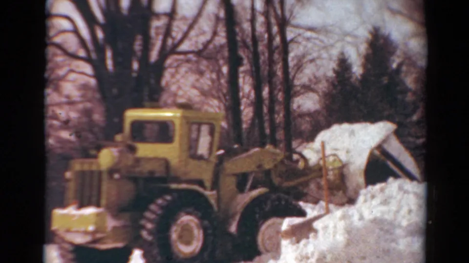 The tractor continues clearing heavy snow near a partly visible house in Toledo, Ohio, with bare winter trees behind it.