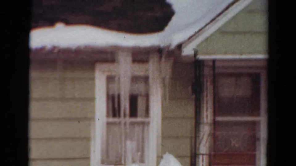1954 Toledo, Ohio 8mm frame of a snowbound residential exterior with white-trimmed windows, pale siding, and a porch or doorway area at the right.