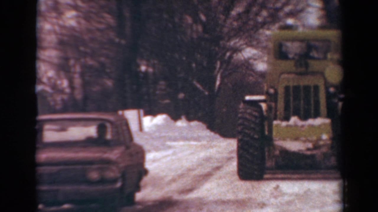 Yellow snowplow approaches on a snowy road as a classic car passes at left.