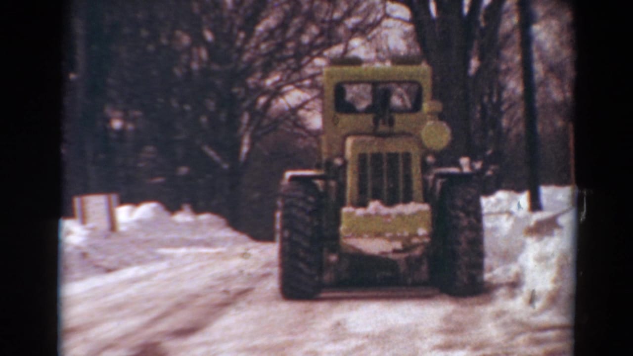 Yellow snowplow drives forward between snowbanks on a tree-lined winter road.