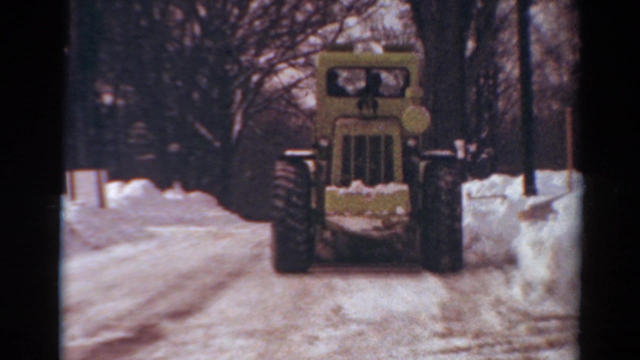 1954 Toledo, Ohio 8mm frame showing the front of a yellow snowplow on a snow-covered street with high snowbanks and bare trees.