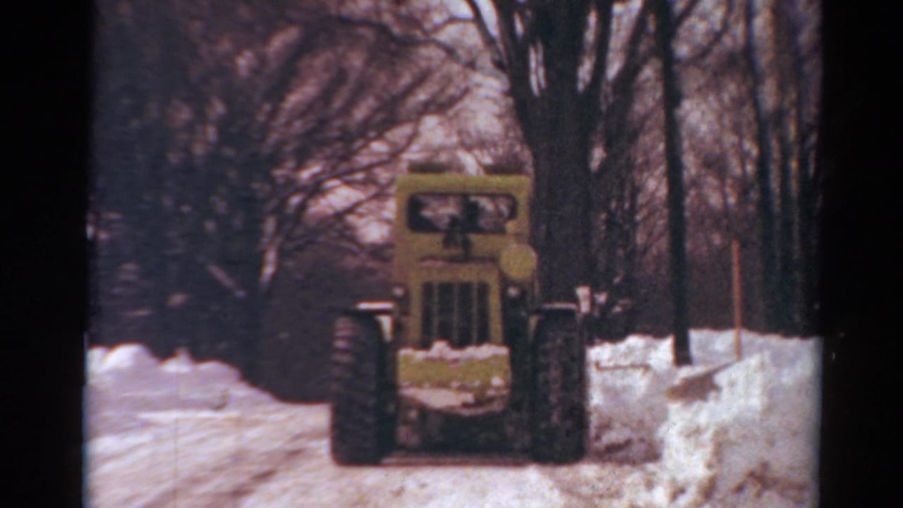 1954 Toledo, Ohio 8mm frame showing a yellow snowplow continuing forward on a snowy road bordered by bare trees and curbside snowbanks.