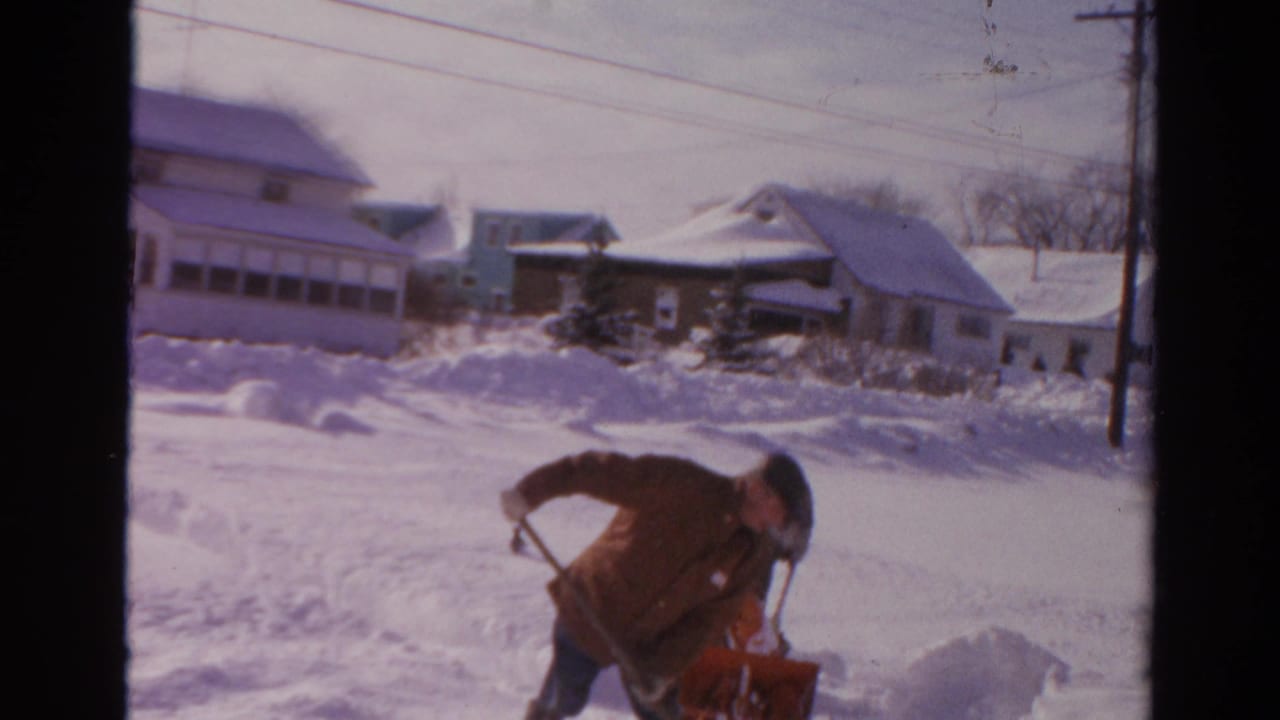 Man in a winter coat bends near a red snow blower on a snowy residential street.