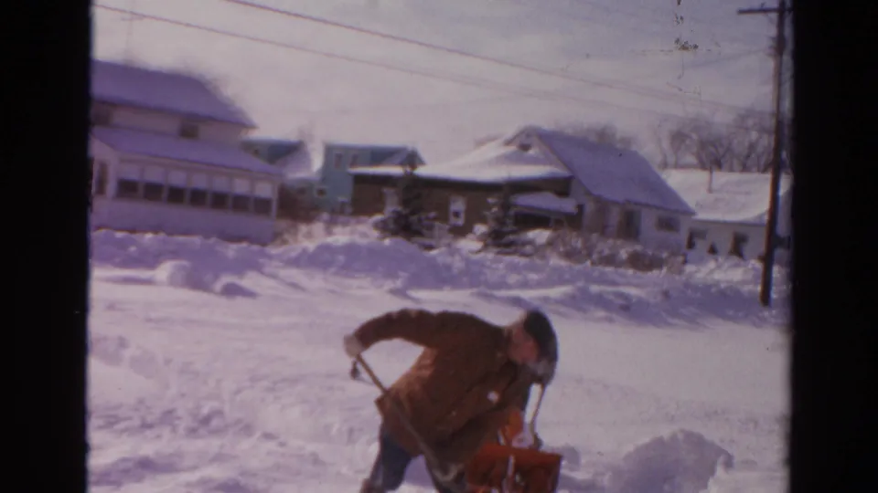 In Toledo, Ohio, 1954 footage, a man works beside a red snow blower amid deep snow and houses in the background.