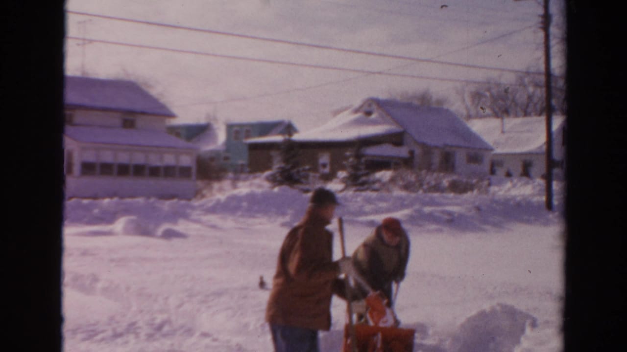 Two men stand by a red snow blower in deep snow with houses behind them.