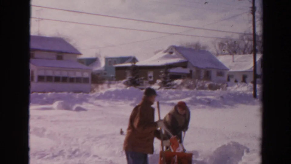 Two men in winter clothing work together near a red snow blower on a snowy Toledo residential street in 1954.