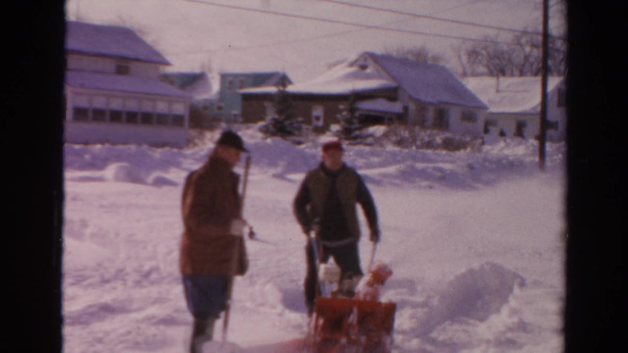 A man with a shovel stands beside another man operating a red snow blower in deep snow.