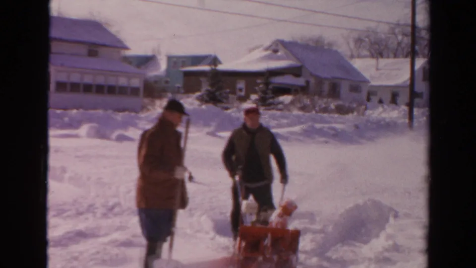 In Toledo, Ohio, two men work in a snowy residential street, one holding a shovel and one guiding a red snow blower.
