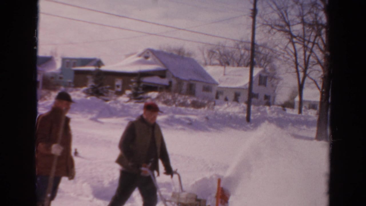 A red snow blower is pushed through deep snow while another man stands nearby with a shovel on a Toledo residential street.