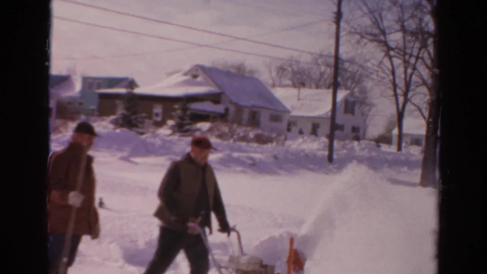 A red snow blower is pushed through deep snow while another man stands nearby with a shovel on a Toledo residential street.