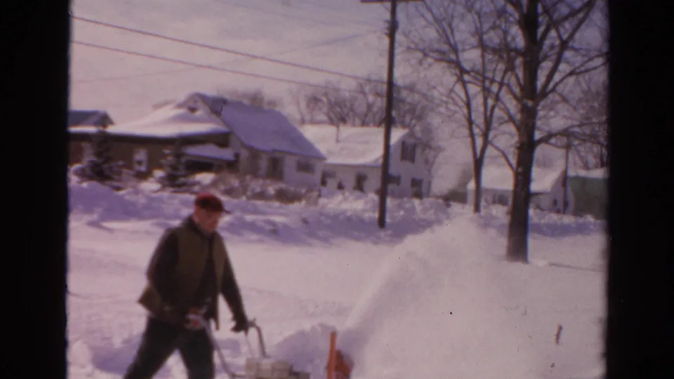 In Toledo, Ohio, 1954 home-movie footage, a man in a red cap operates a snowblower beside snowbanks and houses.