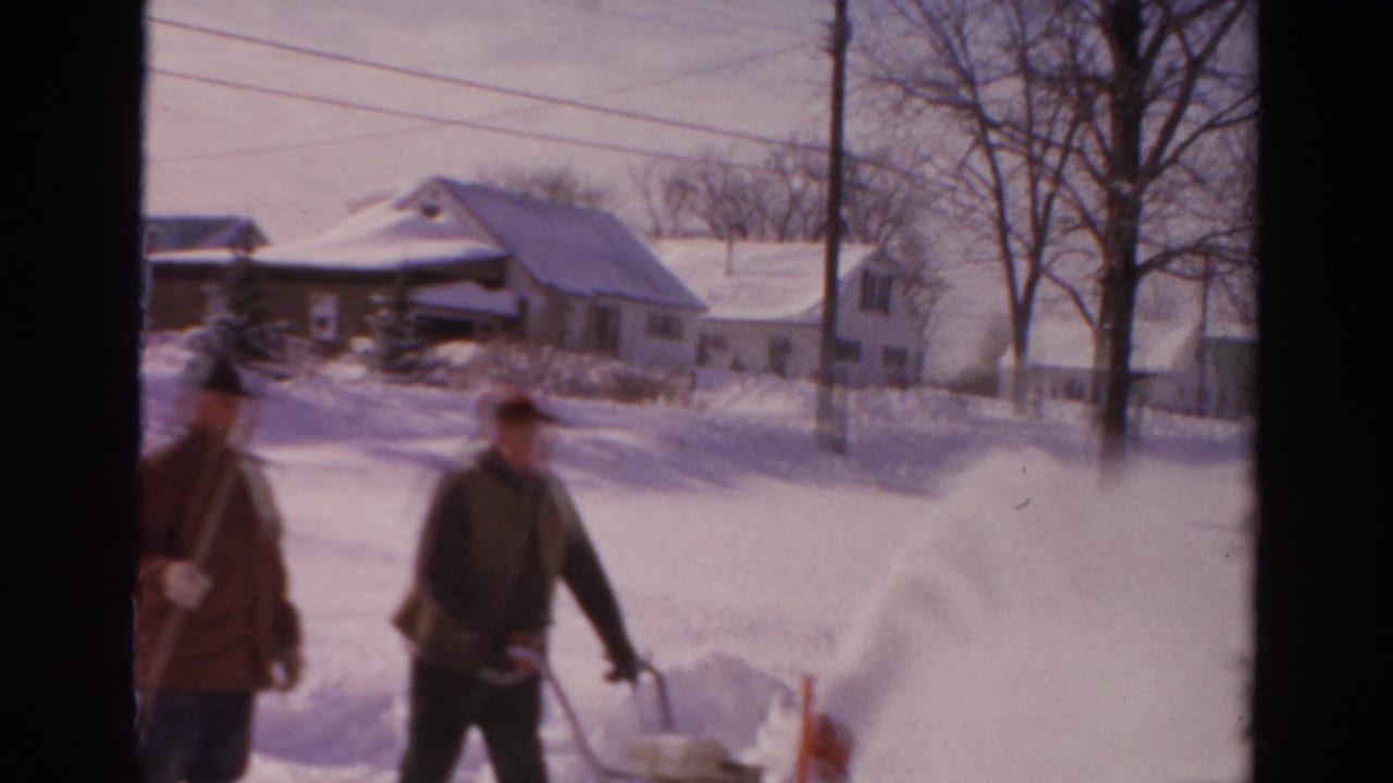 Two men move along a snowy residential street, one pushing a snowblower and one carrying a shovel.