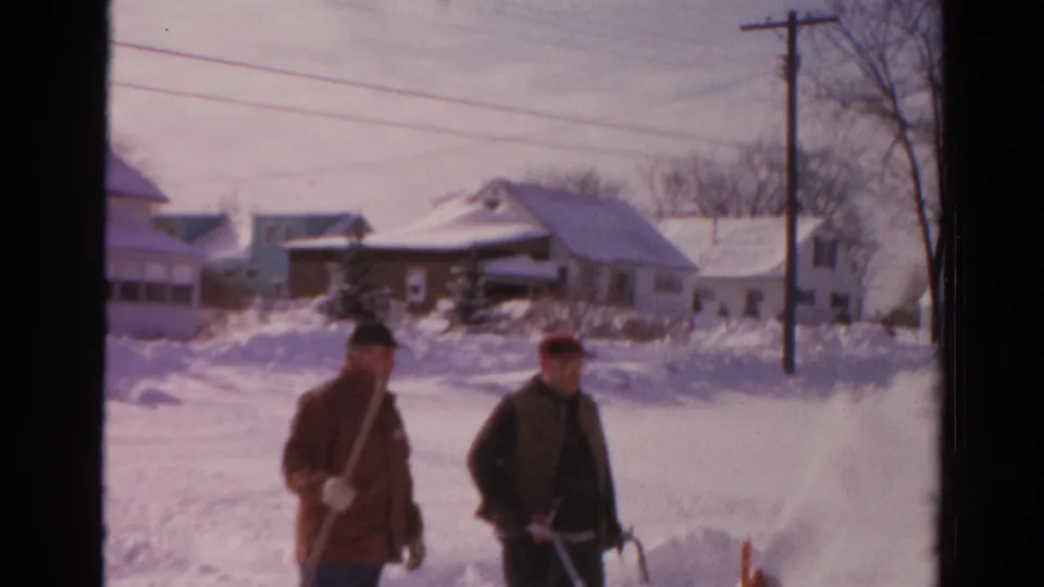 The two men continue past snow-covered homes in Toledo, with a shovel, snowblower, bare trees, and utility lines visible.