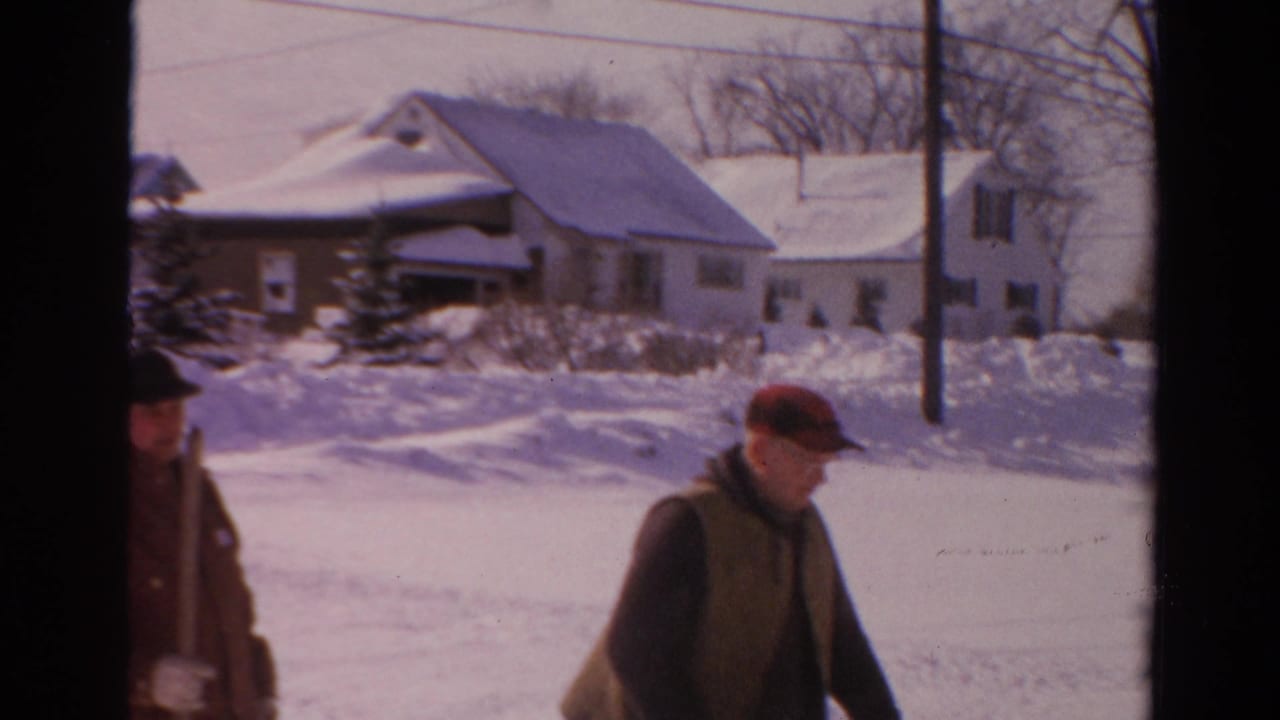 Two bundled men stand in a snowy residential street with houses and snowbanks behind them.