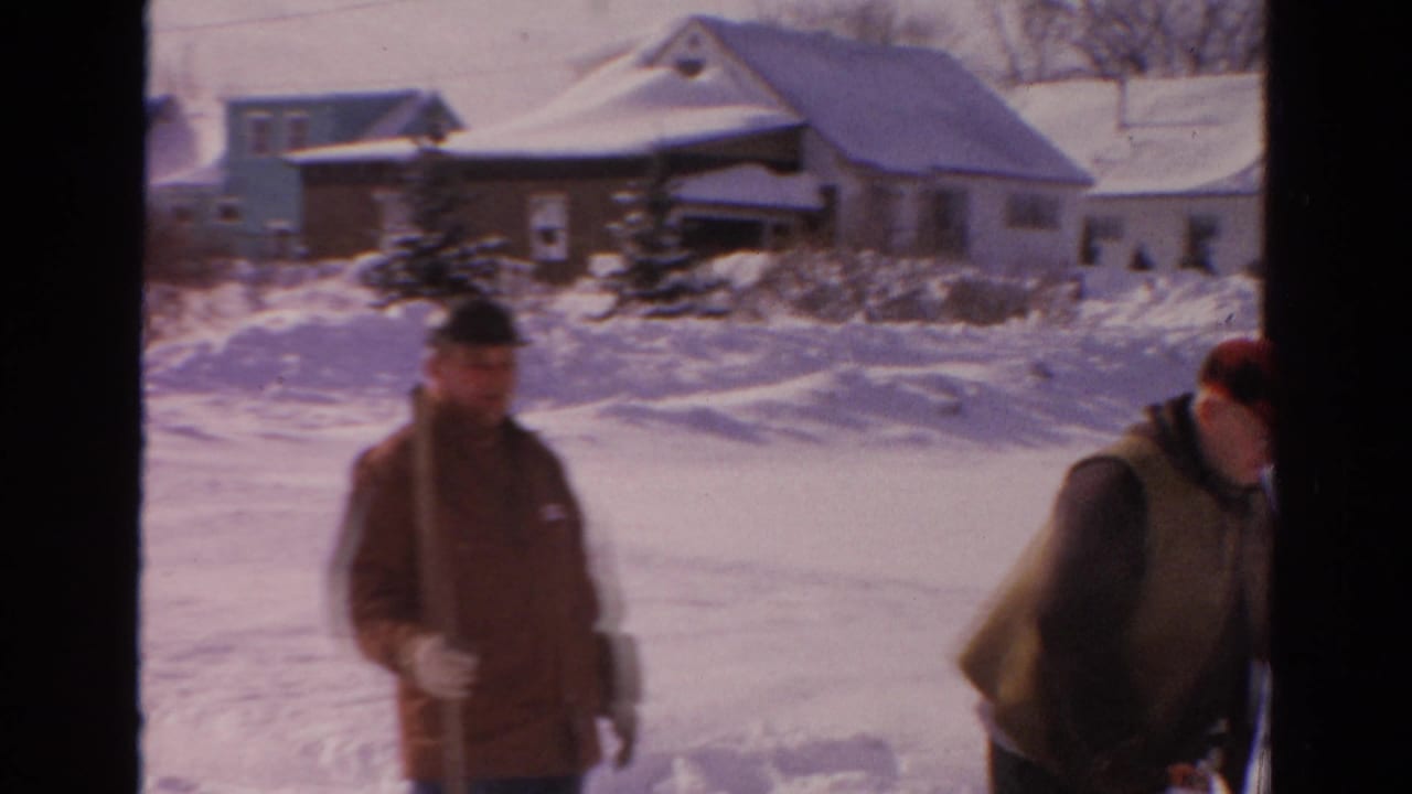 A man in a heavy coat stands in the snowy street while another man is partly visible at the edge.