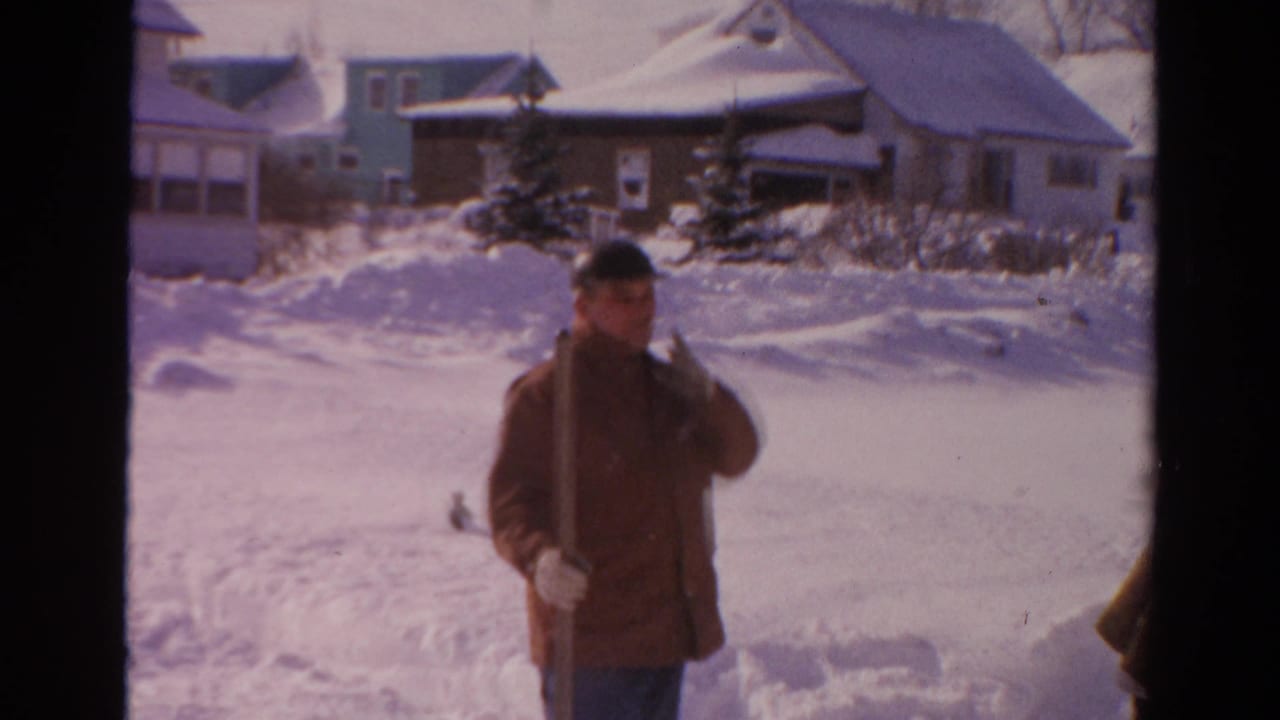 A 1954 8mm frame shows a man in winter clothing pausing in a snow-covered Toledo street with houses behind him.
