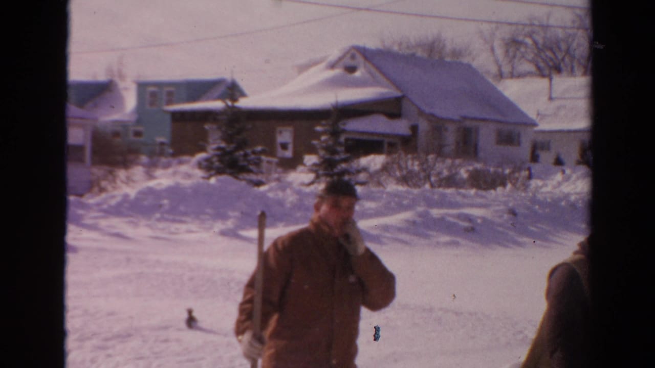A bundled man holding a shovel stands in a snow-covered Toledo neighborhood street, with another person partly at frame edge.