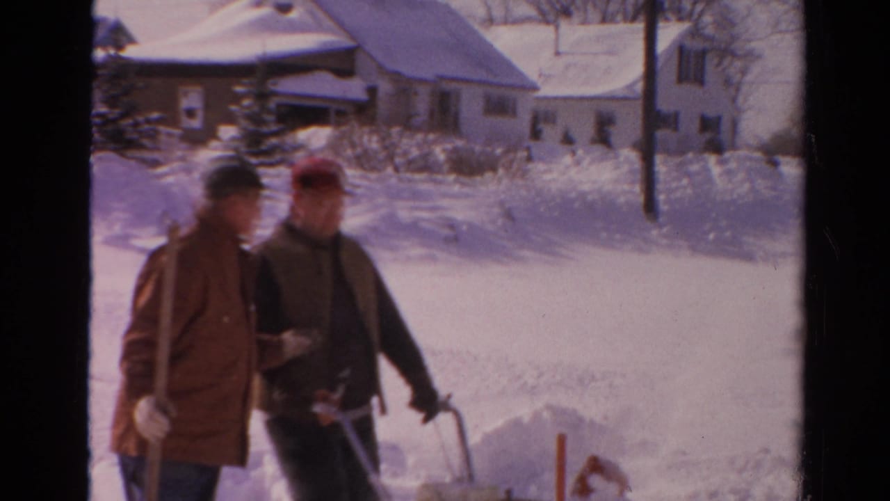 Two adult men in coats and caps stand with snow-removal tools in a Toledo neighborhood yard or sidewalk area.