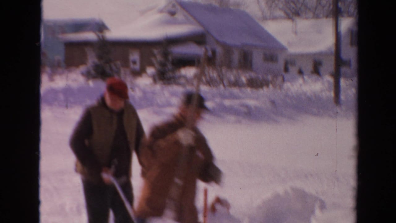 Two adult men move through deep snow with shovels in a residential Toledo winter scene.