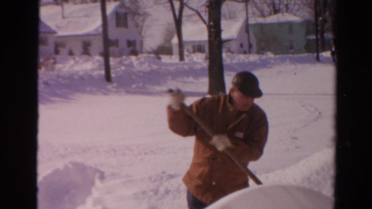 Adult man in a brown winter coat lifts snow with a shovel near a tree and residential houses in Toledo.
