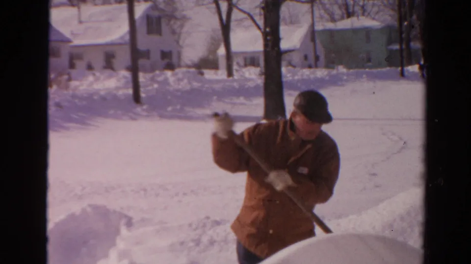 Adult man in a brown winter coat lifts snow with a shovel near a tree and residential houses in Toledo.