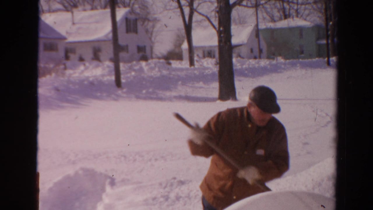 Man in a brown jacket shoveling deep snow near a residential street.