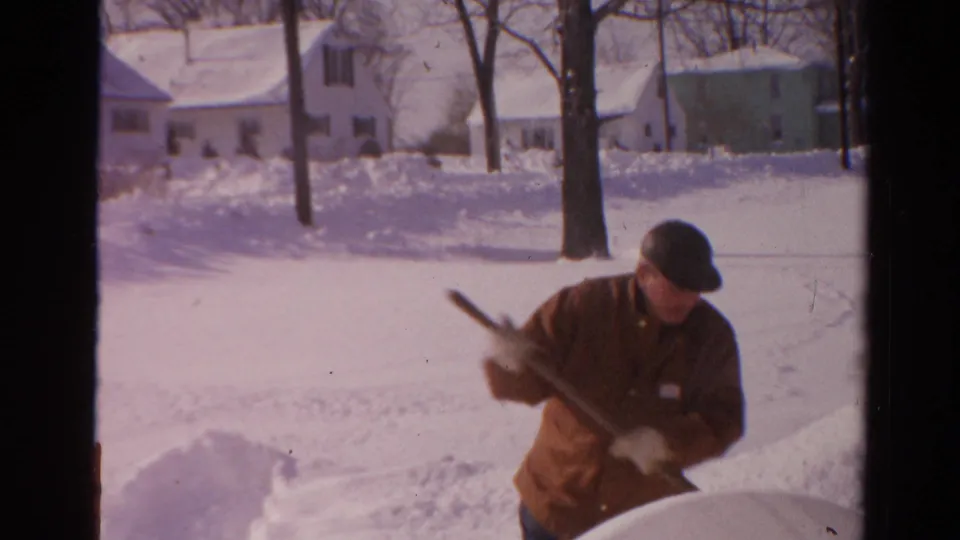 In 1954 Toledo home-movie footage, a man in a brown jacket and dark cap shovels snow with houses and bare trees behind him.
