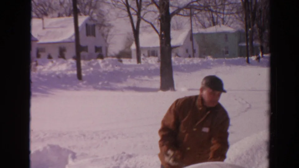The man remains near the snowbank in the Toledo winter scene, with snow-covered lawns, houses, and leafless trees in the background.