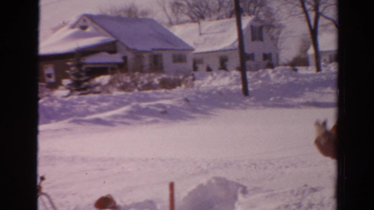 A wider home-movie view shows snow-covered houses and trees in Toledo, with part of the snow-clearing figure entering at the right edge.