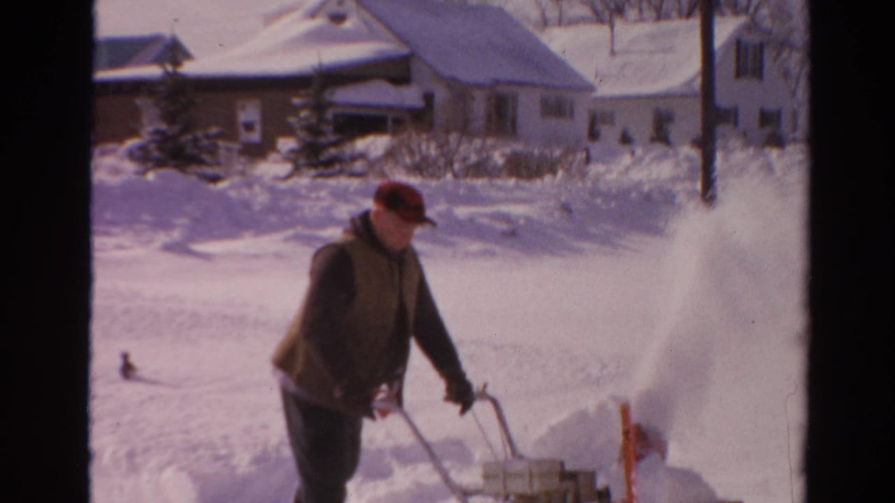 1954 Toledo home-movie frame showing a man in a red cap operating a snow blower near snowbanks and detached houses.